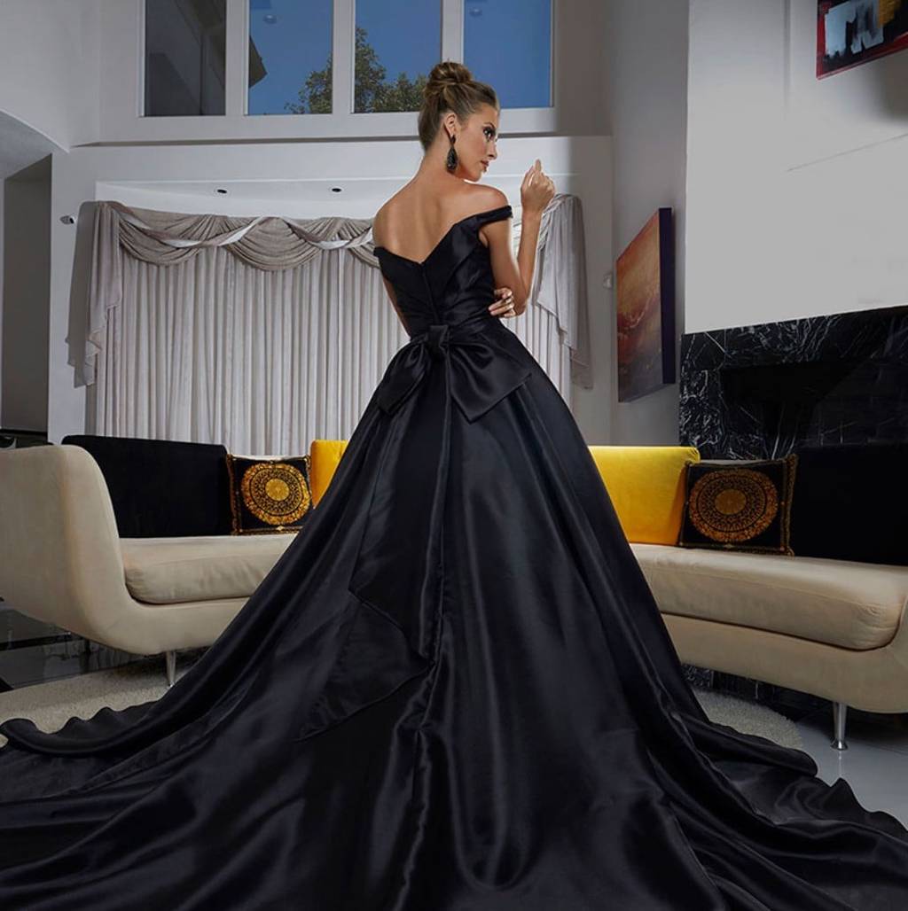 Model seen from behind in a black off-the-shoulder satin ball gown with a bow at the waist and a sweeping train